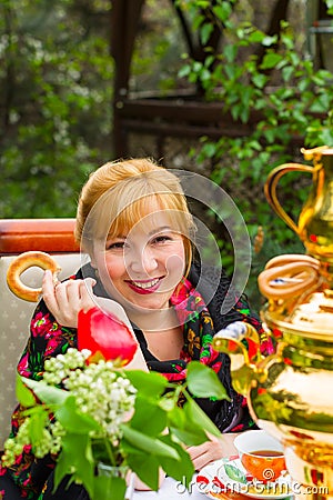 Cheeked Russian cheerful young woman with a scarf draped pavloposadskie, sitting at a table with a samovar and drinking tea with Stock Photo