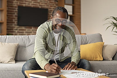 Cheerful adult black businessman in glasses makes notes, studies and works in living room interior, copy space Stock Photo