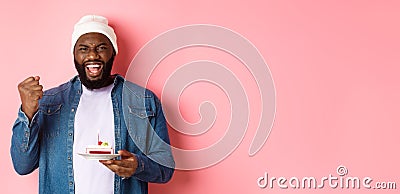 Cheerful african-american guy celebrating birthday, making wish on bday cake with lit candle, smiling happy, standing Stock Photo