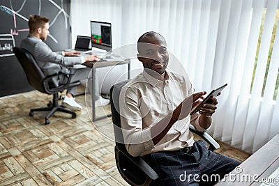 Get your financial attitude high. Cheerful african male trader sitting by desk and studying analytical reports using Stock Photo