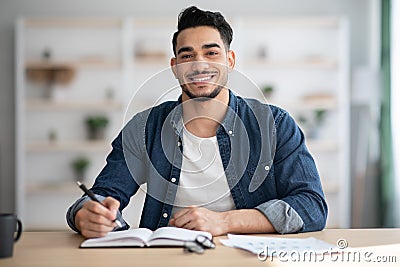 Cheerful arab guy in casual sitting at workdesk, taking notes Stock Photo