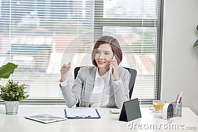 Cheerful asian female talking on mobile phone while sitting on desk with laptop. Business woman in casuals making a phone call and Stock Photo