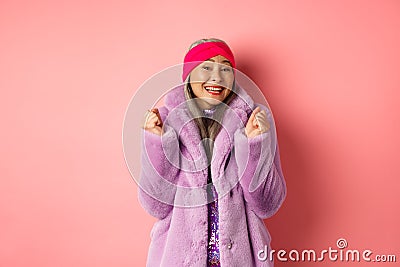 Cheerful asian lady in stylish funky coat, celebrating victory or success, saying yes and smiling happy, standing over Stock Photo