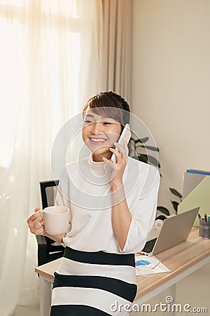 Cheerful Asian woman having conversation on mobile phone and drining coffee at office Stock Photo