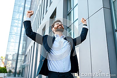 Cheerful attractive young caucasian businessman with beard in suit raises his hands up and rejoices to victory Stock Photo
