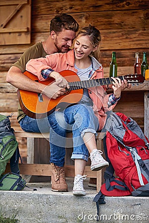 Cheerful attractive young couple enjoying vacation day and playing guitar in front of wooden cabin. Beuatiful moments. Vacation, Stock Photo