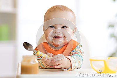 Cheerful baby child eats food itself with spoon. Portrait of happy kid boy in high-chair. Stock Photo