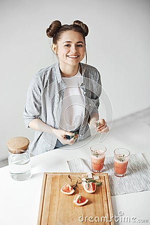 Cheerful beautiful girl smiling decorating grapefruit detox smoothie with rosemary over white wall. Healthy nutrition. Stock Photo