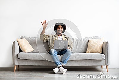 Cheerful Black Guy Using Laptop At Home And Waving Hand At Camera Stock Photo