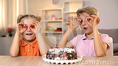 Cheerful brother and sister holding cake cherries front eyes, playing together Stock Photo