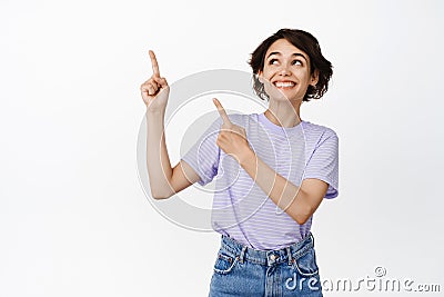 Cheerful brunette girl looks dreamy and happy, pointing fingers at upper left corner, showing banner advertisement Stock Photo