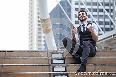 Cheerful Business Man Celebrating Success with mobile phone sitting on the stairs in urban city outdoors, looking up , good news, Stock Photo