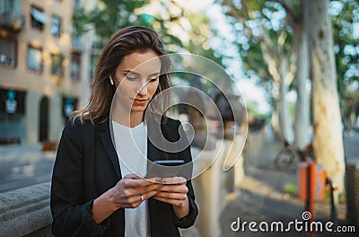 Cheerful businesswoman in jacket with cell phone in park, portrait elegant woman with smartphone and wireless earphones on street Stock Photo