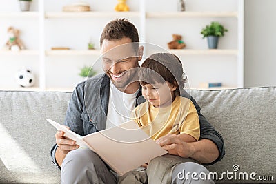 Cheerful carying daddy reading book to little son, baby boy enjoying fairy tale on dad& x27;s knees, sitting on sofa at home Stock Photo