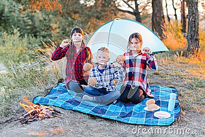 A cheerful company of two girls and a boy on a picnic in the middle of the forest. Children fry sausages on the fire, eat buns and Stock Photo