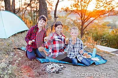 A cheerful company of two girls and a boy on a picnic in the middle of the forest. Children fry sausages on the fire, eat buns and Stock Photo