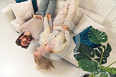 Cheerful couple lying upside-down on couch with colourful cushion in living room Stock Photo