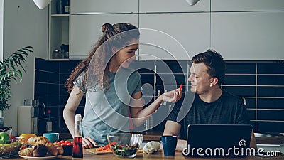 Cheerful couple at table woman cooking while gives hasband to try red pepper, man sitting working on laptop Stock Photo
