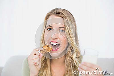 Cheerful cute blonde eating cookie with milk sitting on cosy sofa Stock Photo