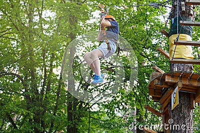 Cheerful cute young boy in blue t shirt and orange helmet in adventure rope park at sunny summer day. Active lifestyle, sport Stock Photo