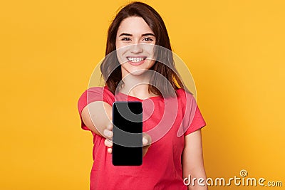 Cheerful energetic black haired female reaching out arm, holding smartphone in one hand, smiling sincerely, looking directly at Stock Photo