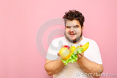 Cheerful fat man in white t-shirt stands on a pink background and holds fruits and salad leaves in his hands, looks in camera and Stock Photo