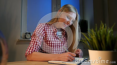 Cheerful female writing in daily planner. Lively young blond woman in casual checkered shirt sitting at wooden desk with Stock Photo