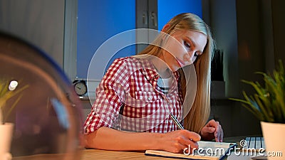 Cheerful female writing in daily planner. Lively young blond woman in casual checkered shirt sitting at wooden desk with Stock Photo