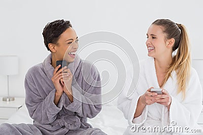 Cheerful friends in bathrobes text messaging on bed Stock Photo