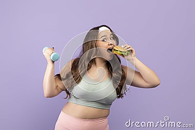 Cheerful funny hungry pretty millennial plus size young female in sports uniform eating burger and lifting dumbbell Stock Photo