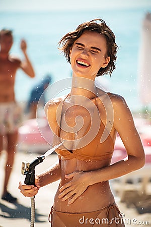 Cheerful girl in bikini using hose pipe on beach. Portrait of smiling lady in beige swimsuit rinsing beach sand off her Stock Photo