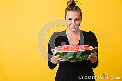 Cheerful girl in black dress joyfully looking in camera while holding half of big watermelon in hands over yellow Stock Photo