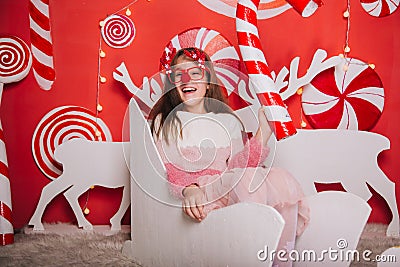 A cheerful girl in glasses with a red nose and horns is holding a candy in her hands. A wall of lollipops. The child is sitting in Stock Photo