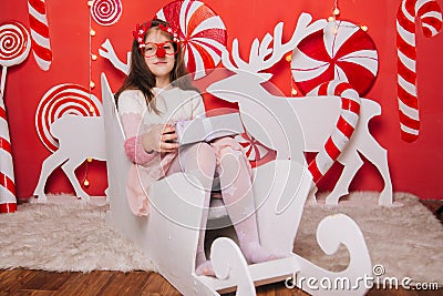 A cheerful girl in glasses with a red nose and horns is holding a gift in her hands. A wall of lollipops. The child is sitting in Stock Photo