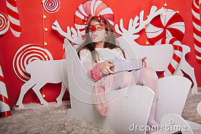 A cheerful girl in glasses with a red nose and horns is holding a gift in her hands. A wall of lollipops. The child is sitting in Stock Photo