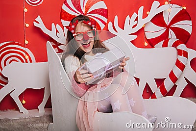 A cheerful girl in glasses with a red nose and horns is holding a gift in her hands. A wall of lollipops. The child is sitting in Stock Photo