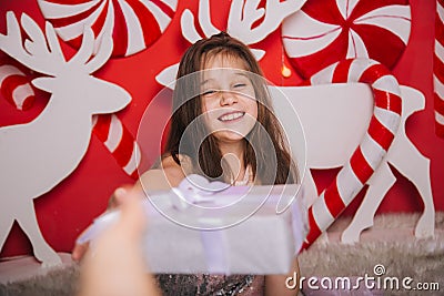 Cheerful girl holding a Christmas present in her hands. A wall of lollipops. A child sits on a carpet against the background of Stock Photo