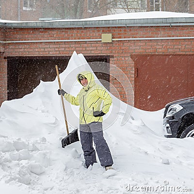Cheerful girl with the shovel for snow removal stands near a huge snowdrift near the garage Stock Photo