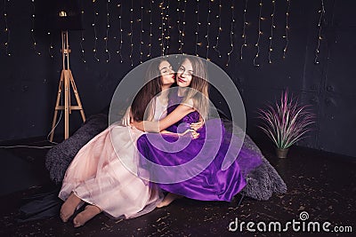 Cheerful girlfriends. Two long-haired girls in pink and purple dresses in studio on dark background with bokeh. Copy space. Stock Photo