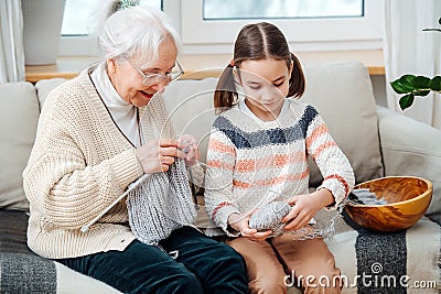 Cheerful granny knitting on a couch at home, her granddaughter helping her Stock Photo