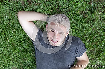 A cheerful guy in a gray T-shirt is lying on a green lawn with one eye squinted Stock Photo