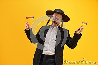 Cheerful Jewish man in hat, with sidelocks holding noisemaker, against yellow background. Celebration Stock Photo