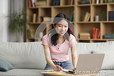 Cheerful korean lady attending webinar from home, sitting on sofa, using laptop and taking notes, copy space Stock Photo