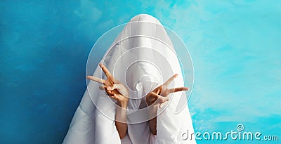 Cheerful lazy woman waking up after sleeping covered head with white blanket showing cool gesture her fingers on blue background Stock Photo