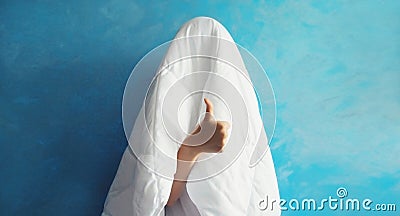 Cheerful lazy woman waking up after sleeping covered head with white blanket showing cool gesture her fingers on blue background Stock Photo
