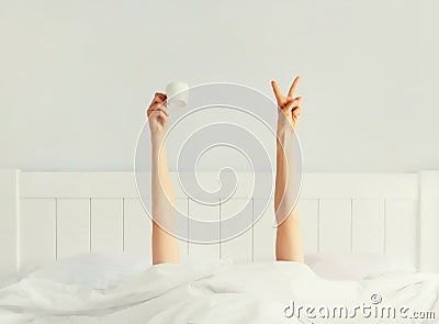 Cheerful lazy woman waking up after sleeping lying in soft comfortable bed showing empty cup coffee stretching her hands up from Stock Photo