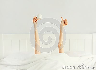 Cheerful lazy woman waking up after sleeping lying in soft comfortable bed showing empty cup coffee stretching her hands up from Stock Photo