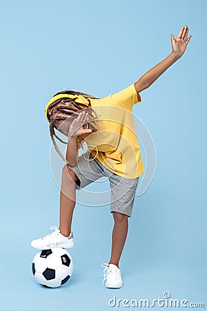 Cheerful little boy with african with headphones dabbing with one leg on soccer ball over blue background. Stock Photo