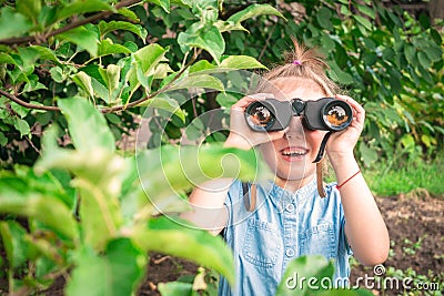 A cheerful little girl is looking through binoculars from the bushes. The child is watching the parents at a distance. The baby Stock Photo