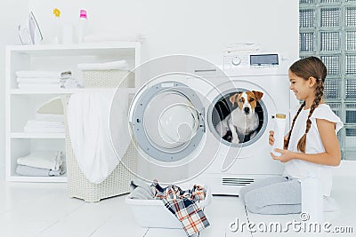 Cheerful little girl stands on knees with washing detergent, poses near washing machine, looks happily at favourite pet inside of Stock Photo